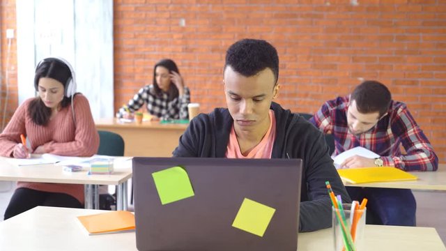 young attractive man sitting at desk in office working behind laptop. looks pleasantly into the camera with a smile. in the background a large office with tables and people of a mixed race. 4k