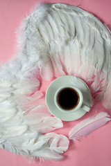 Cup of coffee on a white saucer. White wings of an angel  on a pastel pink background. Feathers....