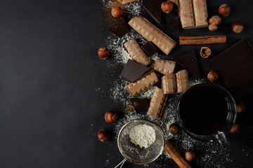 A cup of coffee on a black background. Pieces of milk and dark chocolate, hazelnuts, cinnamon and powdered sugar on a dark background. Flat lay. Copy spase