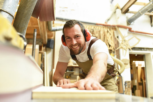 fröhlicher Tischler schleift Holzbrett an einer Maschine in Schreinerei // carpenter works in a joinery - workshop for woodworking and sawing