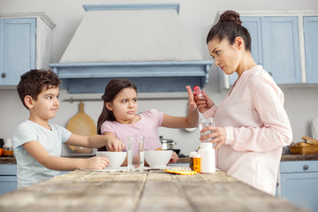 Oh no. Pretty annoyed dark-haired little girl having breakfast with her brother and refusing to take vitamins which her mom giving her