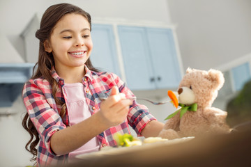 So tasteful. Pretty content dark-haired little girl smiling and feeding her toy with healthy food while sitting at the table in the kitchen