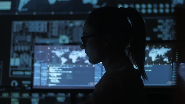 silhouette of young woman IT programmer working at a computer in the data center filled with display screens