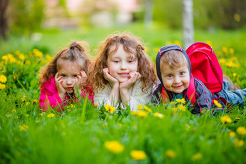Three children lie on the green grass with yellow dandelions. Two sisters and a brother in the street. The boy is smiling.