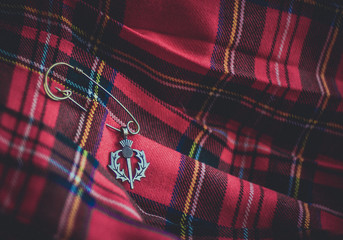 A vintage brass thistle pendant on a pin against the background of a traditional scottish kilt. Symbols of Scotland. Selective focus. - obrazy, fototapety, plakaty