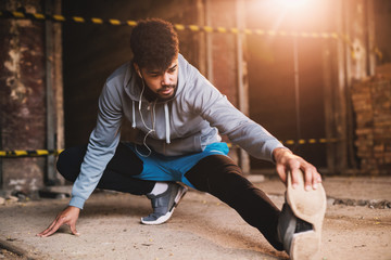 Naklejka premium Close up portrait of active afro-american young attractive athletic man doing full leg stretching workout inside of the abandoned place.