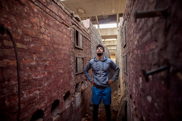 Obraz na płótnie Canvas Portrait of focused motivated afro-american young handsome sportive man with earphones standing inside of the abandoned place in the middle of two walls and thinking while looking up.