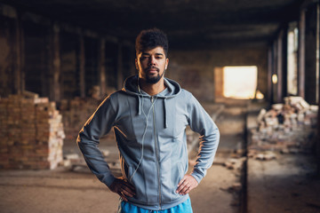 Portrait of focused motivated afro-american young handsome sportive man with earphones standing inside of the abandoned place. Successful young active man portrait.