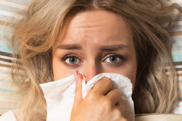 Closeup Of Beautiful young Caucasian upset and sick female with paper napkin blowing nose, lies on the pillow, looking at camera, indoor/ Rhinitis, cold, sickness, allergy concept