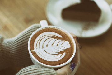 Top view female hands holding a cup of hot coffee with heart shaped latte art and plate of chocolate cake on the table.