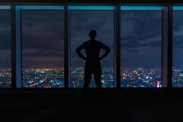 Fototapeta na wymiar Man looking out large windows high above a sprawling city at night