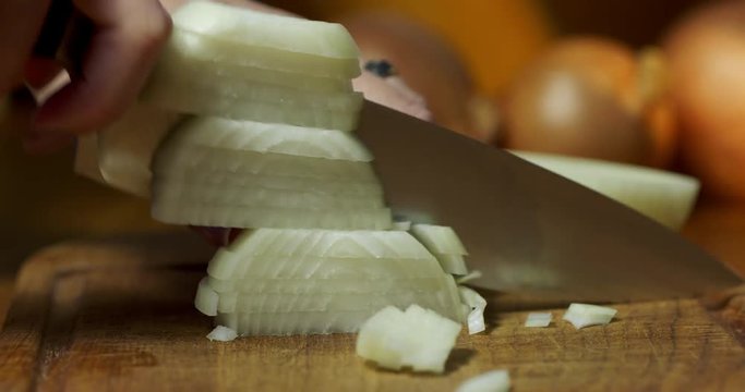Woman's hand chops up onion with large knife on wooden board in kitchen. Recorded in 4K with dolly move.