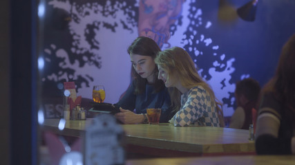 Two beautiful young women sitting at cafe and looking at digital tablet. Teamwork, two young businesswomen sitting at table in cafe. An attractive woman and her friend hold a tablet