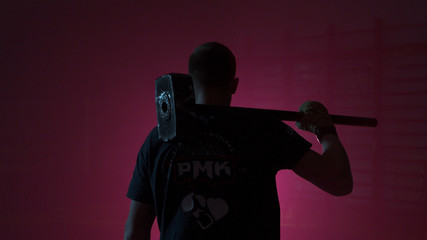 Man with a sledgehammer behind his back. Guy with a sledgehammer in the red room. Man with a big hammer or a sledgehammer behind comes into the room with a red light. Muscular man with hammer from - obrazy, fototapety, plakaty