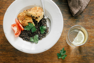 Vegetarian dinner table. Plate with black quinoa and oatmeal cutlets with prunes on wooden table with glass water with lemon, top view