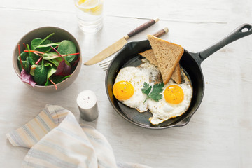Fried egg in frying pan on white wooden table with salad and lemon water, top view