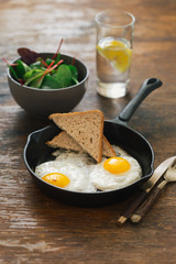Fried egg in frying pan on dark wooden table with salad and lemon water