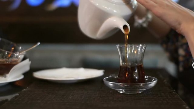 womens hands in a dress and metal watches pour black tea from a white porcelain teapot