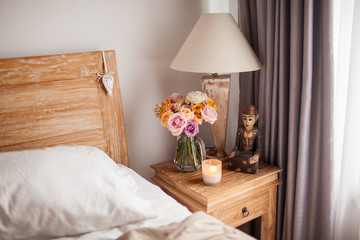 Wooden Bed with White Sheets. A Bedside Table by the Bed with a Lamp, a Bouquet of Flowers from Roses, Candle and Hand Cream