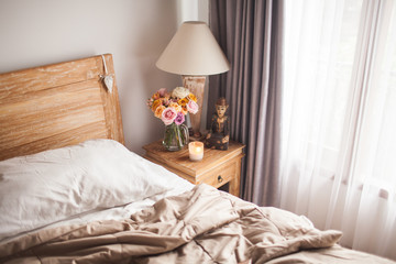 Fototapeta na wymiar Wooden Bed with White Sheets. A Bedside Table by the Bed with a Lamp, a Bouquet of Flowers from Roses, Candle and Hand Cream