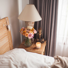 Wooden Bed with White Sheets. A Bedside Table by the Bed with a Lamp, a Bouquet of Flowers from Roses, Candle and Hand Cream