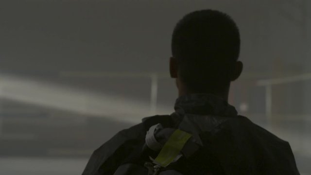Back view on man with a backpack walking in an abandoned old warehouse and light from roof. Guy with a backpack comes into the old sports hall where smoke and light from rooftop. Young man on the old