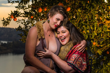 Two young caucasian women dressed in fantasy clothing pose together under an orange tree at sunset