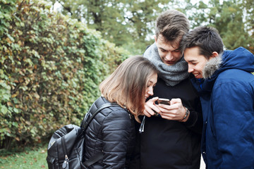 Group of friends two men and one woman using a smartphone in the autumn park . Fall park, three teenage friends look into their phone and laughing.