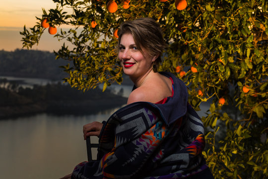 Young caucasian woman with short hair and flashy jacket poses under an orange tree at sunset