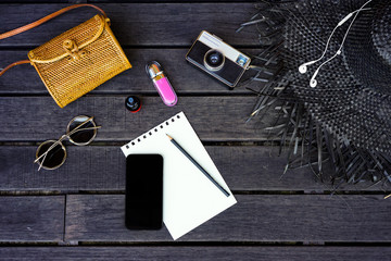 Minimal styled flat lay isolated on dark wood background with digital phone. Woman desk top view with accessories: hat, handbag, sunglasses, vintage photo camera, headphones, notebook, cosmetics