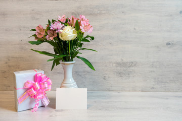 Colorful spring bouquet of rose, chrysanthemum and alstroemeria flowers in a vase with empty card and gift box on wooden background