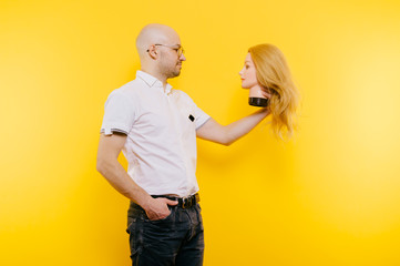 Excited odd bizarre strange unusual  adult bald hairless man holding plastic blonde woman head. Funny male with girlfriend isolated on yellow. Baldness after chemotherapy. Cancer hair loss concept.