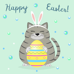 A gray cat in the rabbit's ears sits and holds a yellow Easter egg. Happy easter.