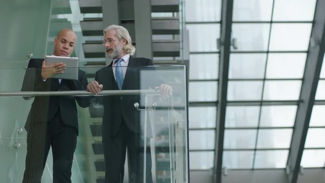 two corporate executives standing on second floor on a glass and steel building discussing business using digital tablet.