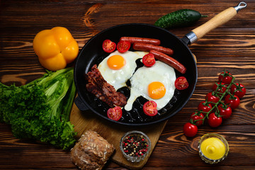 Fried eggs with bacon and sausages in a frying pan with spices and fresh vegetables.