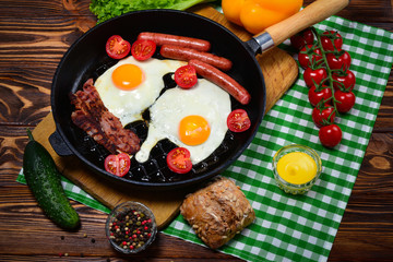 Fried eggs with bacon and sausages in a frying pan with spices and fresh vegetables.