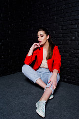 Stylish brunette girl on red jacket against studio black brick wall.