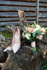 Close up of beautiful wedding bouquet of pink and white roses and beige shiny bride shoes on wooden stump outdoors. Wedding morning preparation. Wedding bride accessories. Bridal bouquet and shoes