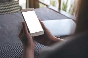 Mockup image of hands holding white mobile phone with blank desktop screen and laptop on table in cafe