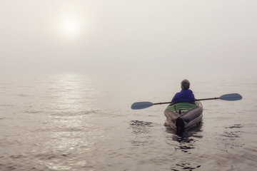Fototapeta na wymiar Girl kayaking on an inflatable kayak in Howe Sound during a fog covered winter sunset. Taken in West Vancouver, BC, Canada.