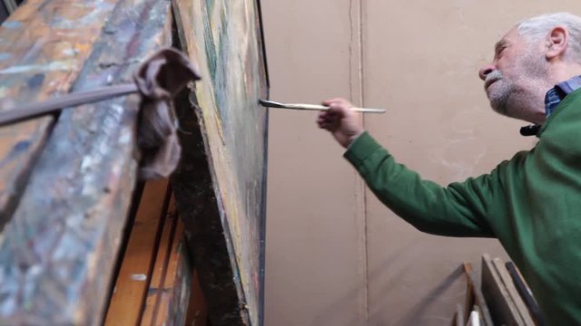 The old artist paints his painting in a workshop of oil painting