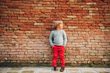Outdoor portrait of handsome little boy wearing red chinos and light blue pullover, stylish kid posing against vintage brick wall. Fashion for small children