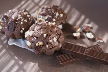 Three chocolate muffins with hazelnuts  and two pieces of chocolate on brown background with the shadow from the shutters in sunny day