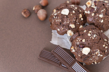 Three chocolate muffins with hazelnuts on the baking paper and two pieces of chocolate near on brown background 