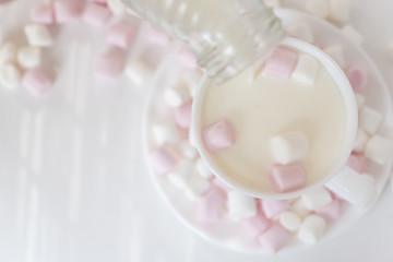 White cup and saucer with white and pink mini marshmallows and a bottle of milk on white background. Milk is pouring into the cup.