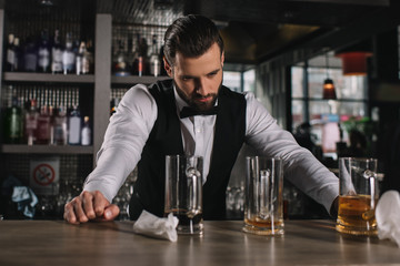 tired handsome bartender leaning on bar counter and looking at dirty glasses and trash