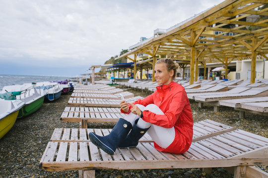 young woman in a red raincoat and blue rubber boots sitting on a wooden deck chair by the stormy sea and sending a text message from her cell phone. Woman using smart phone outdoor