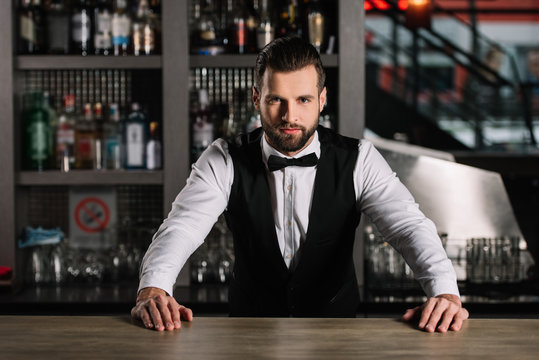 handsome bartender leaning on bar counter and looking at camera