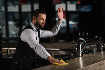 handsome bartender cleaning bar counter and waving hand to someone