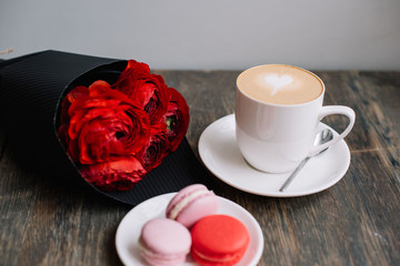 Delicious fresh morning cappuccino coffee with french macarons cookies of red an pink color and red ranunculus flower bouquet on the rustic wooden table background, festive  romantic setup